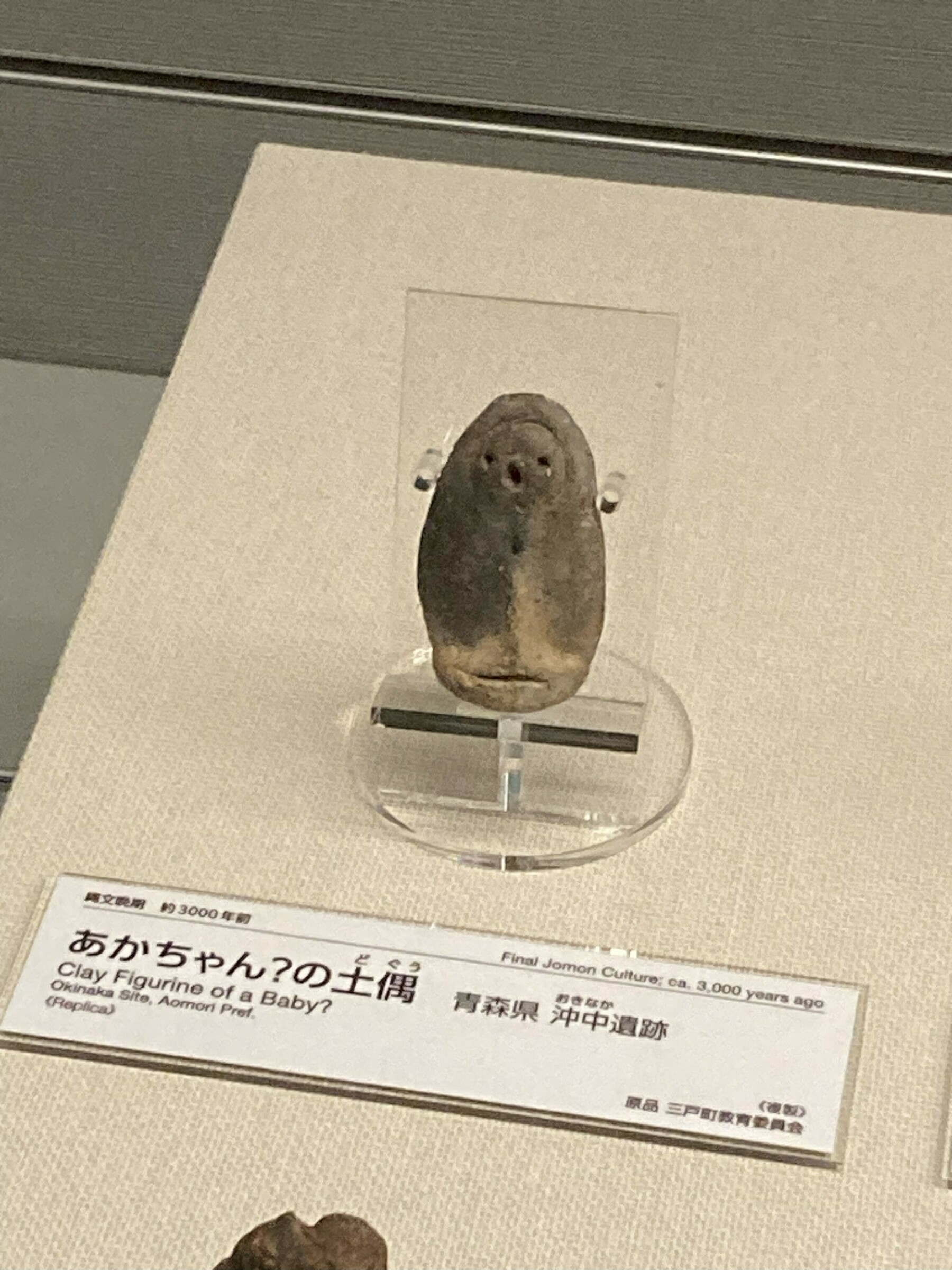 Auto-generated description: ガラスケース内に展示された、顔のような形の土偶が見える。