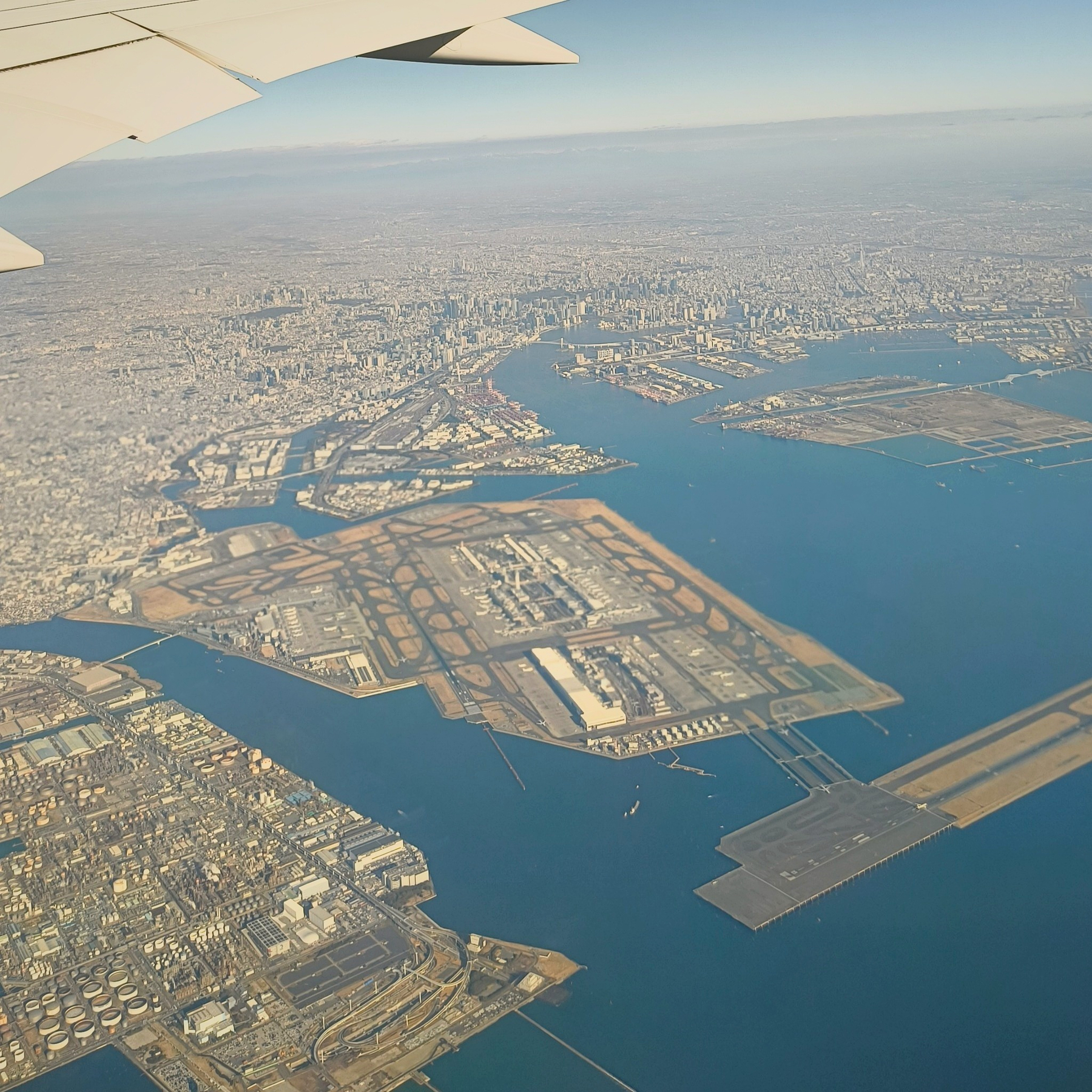 Auto-generated description: 飛行機の翼越しに広大な都市と海が見下ろせる風景。