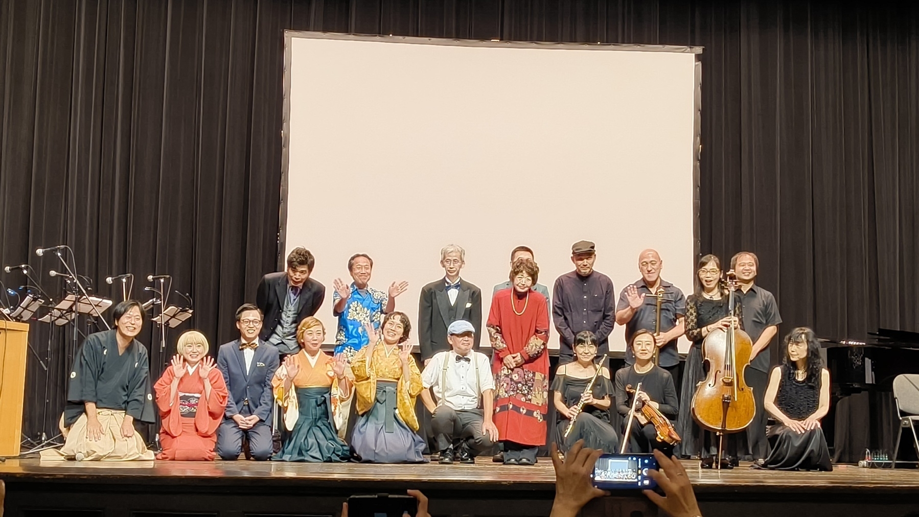 Auto-generated description: 大勢の人々が舞台で並んでポーズをとっており、楽器を持っている人もいます。