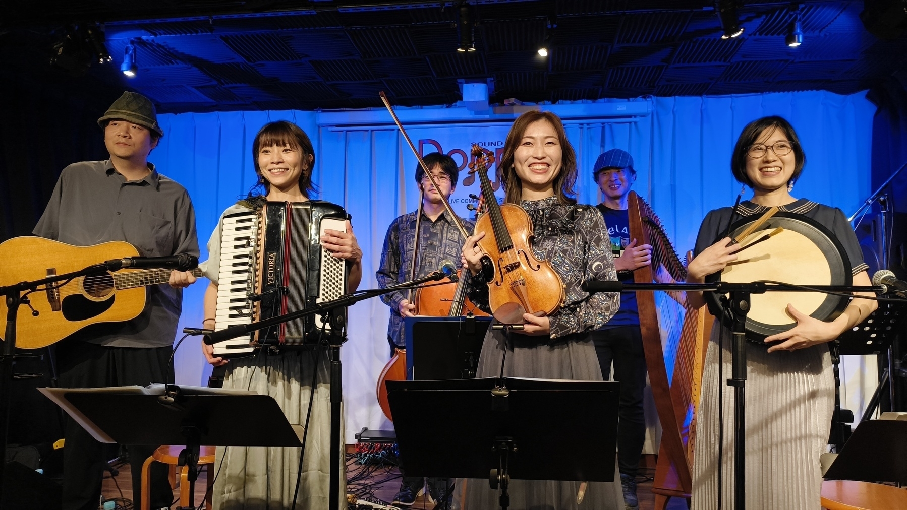 Auto-generated description: 音楽グループがステージ上で楽器を持って微笑んでいる。