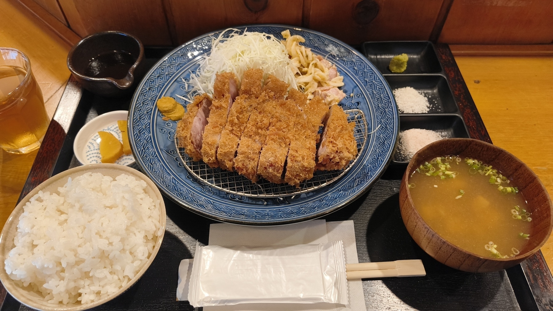 Auto-generated description: カツ定食が盛り付けられた皿が見え、ご飯、味噌汁、キャベツの千切り、お漬物、そして調味料小皿が添えられています。