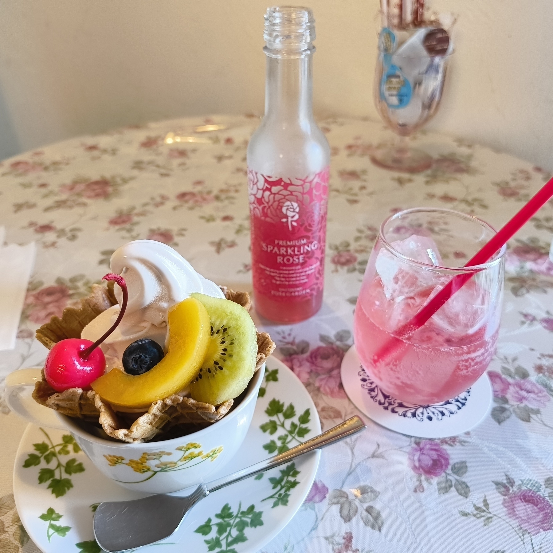 Auto-generated description: 花柄のテーブルに、アイスクリームが乗ったワッフルボウル、フルーツ、チェリー、ソーダとピンク色の飲み物が置かれています。