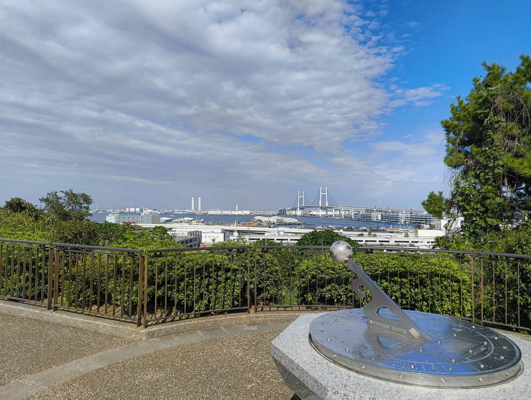 Auto-generated description: 青空の下、緑に囲まれた日の出町公園から横浜ベイブリッジが遠くに見える景色。