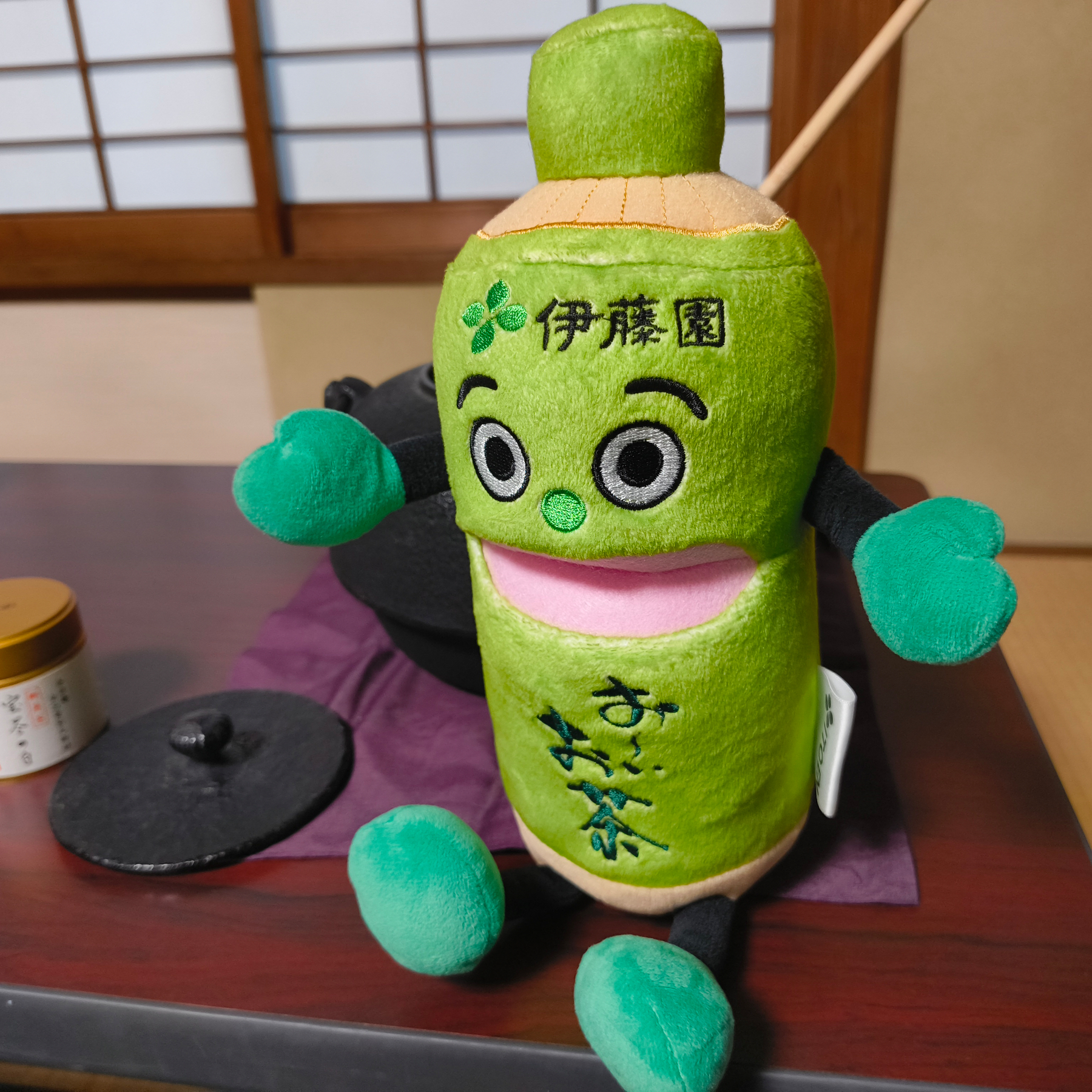 Auto-generated description: 緑茶のボトルを模したかわいいぬいぐるみが畳の部屋の机の上に置かれています。