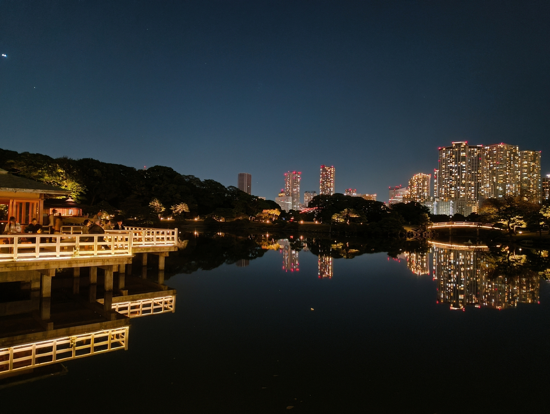 Auto-generated description: 夜景の中、湖に映る都市のビル群と伝統的な建物が見える。