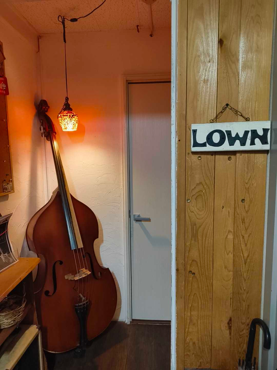 Auto-generated description: コントラバスが置かれた部屋の隅にオレンジ色のランプが灯っており、「LOWN」と書かれたサインが壁に掛かっています。