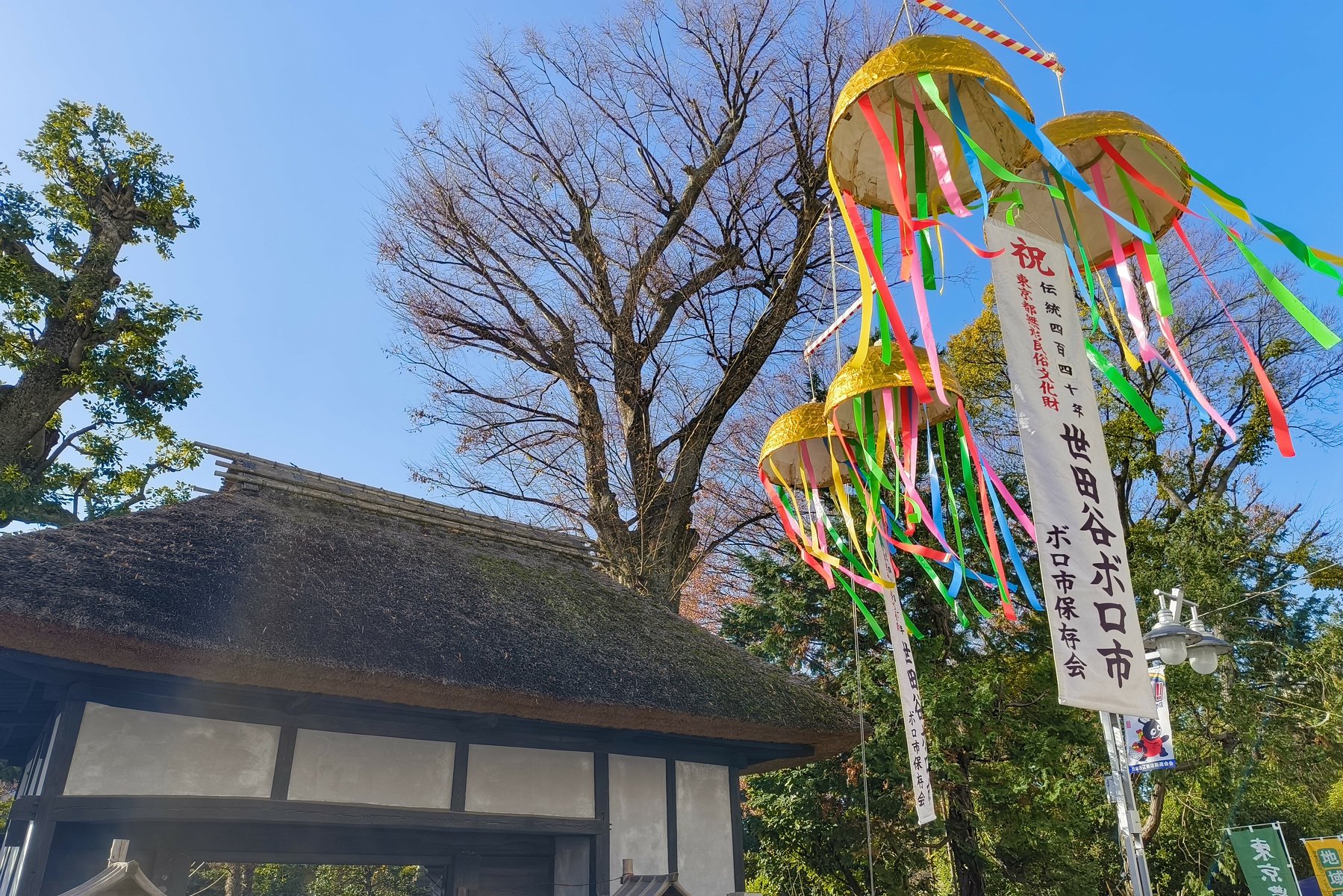 Auto-generated description: 日本の伝統的な藁ぶき屋根の門の近くに、色とりどりのリボンが飾られた吹き流しが立っています。