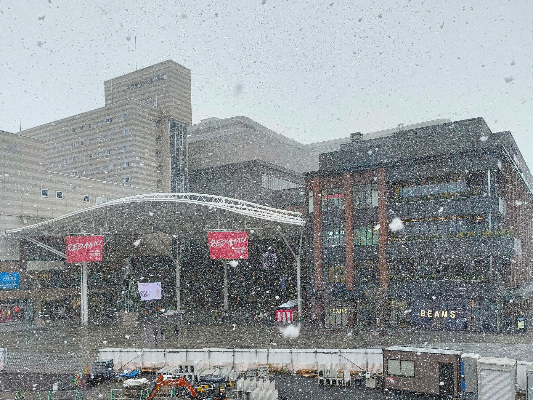 Auto-generated description: 雪が降る中、現代的な建物の前に広場が広がっている都市の風景。