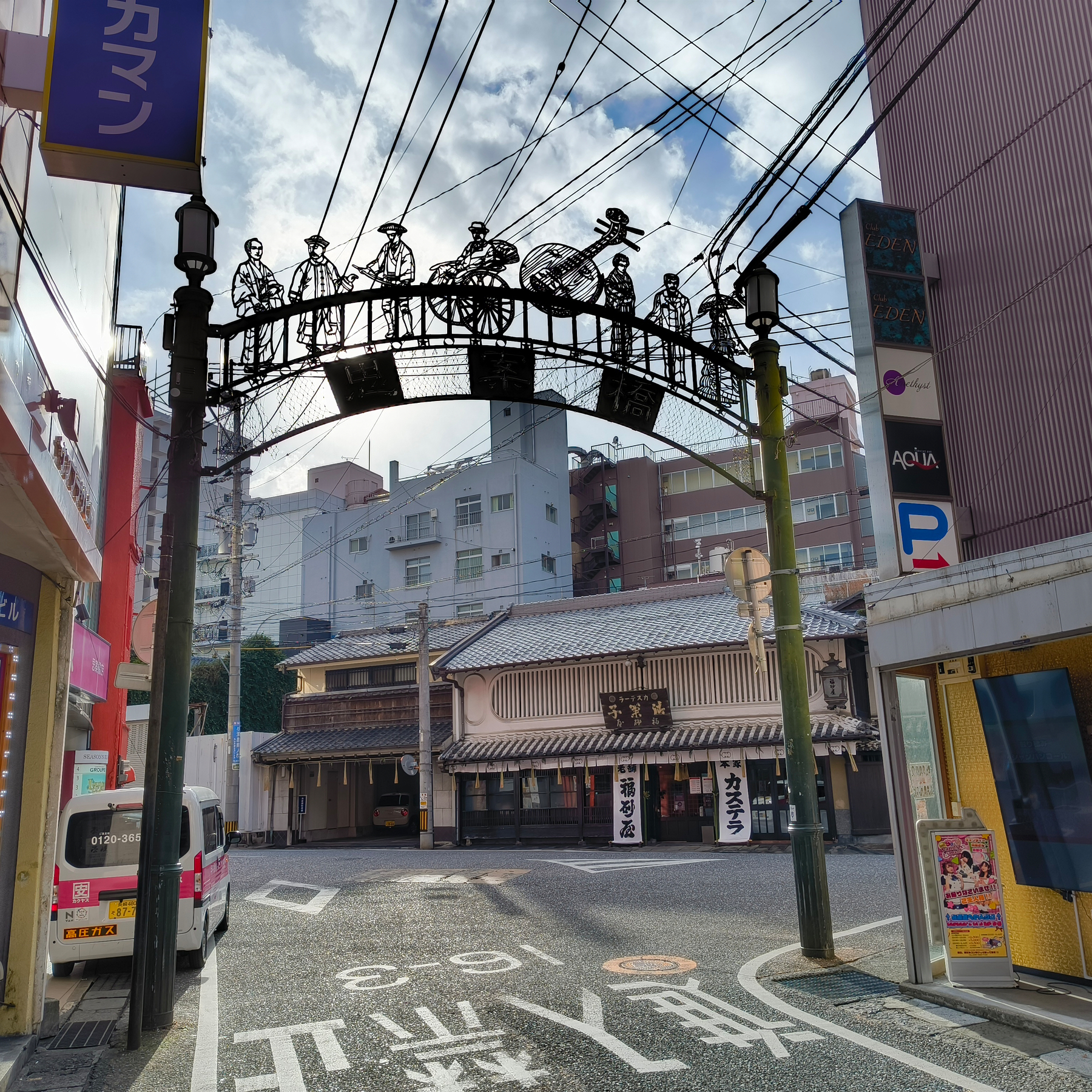 Auto-generated description: 電線と装飾的なアーチが上に架かる、商店が並ぶ通りの風景です。