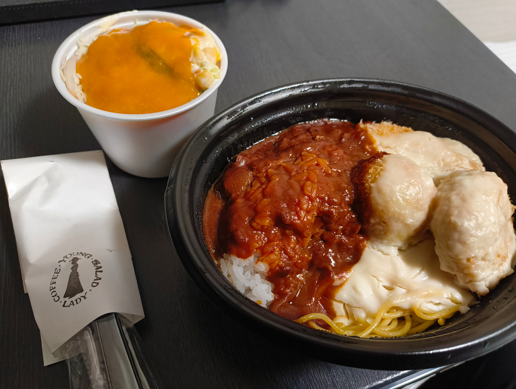 Auto-generated description: カレーとライスが入ったボウルの横には、サラダカップが置かれており、スプーンとフォークが包まれた紙ナプキンが添えられています。