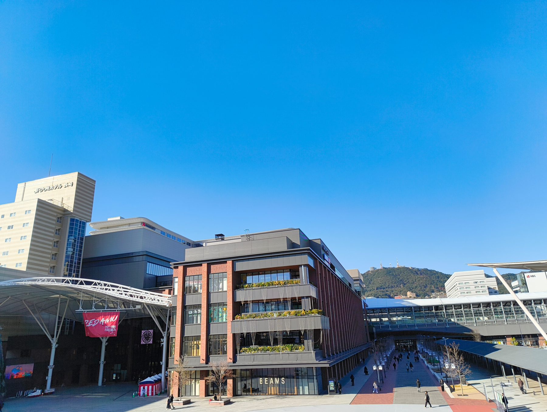 Auto-generated description: 広場に面した現代的なビルと、快晴の空が広がる街の風景。