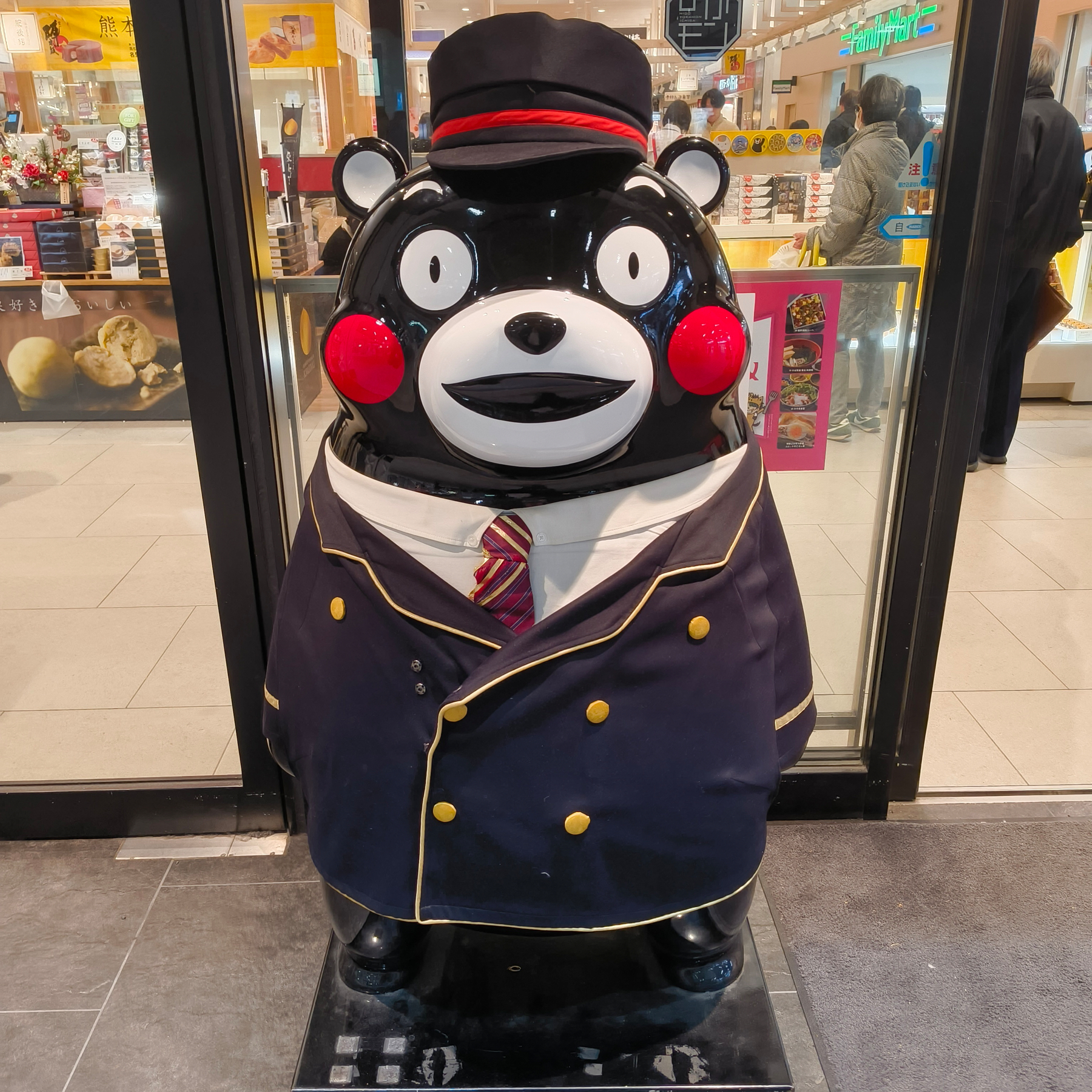 Auto-generated description: クマをモチーフにしたキャラクターが、制服を着た状態で商業施設の入り口に立っています。