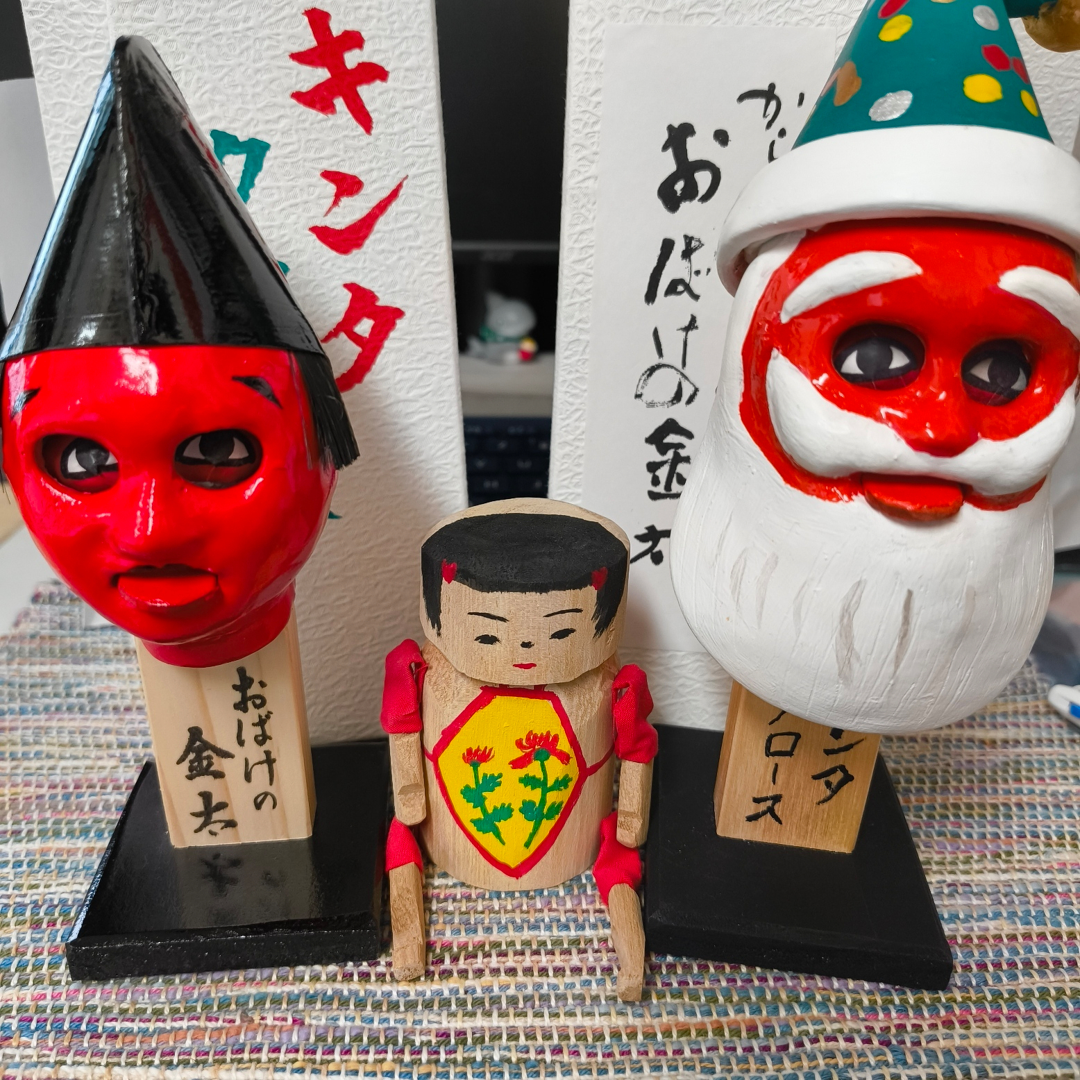 Auto-generated description: 赤い顔のお面と白いヒゲのサンタクロースの人形、中央には絵柄のある小さな木の人形が並んでいます。