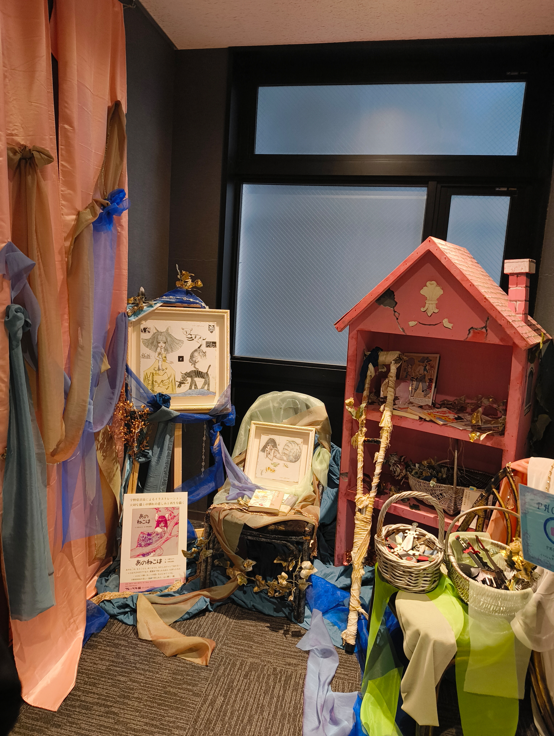 Auto-generated description: 部屋の一角に、絵画や小さな赤い屋根の模型、布で装飾された展示物が配置されています。