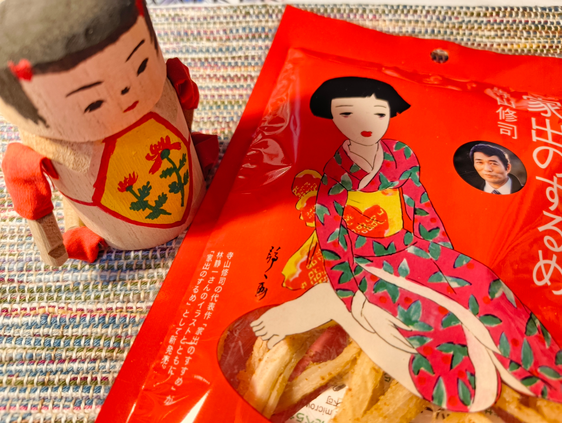 Auto-generated description: 赤い着物を着た女性が描かれたパッケージのお菓子の横に、同じ和風のデザインを持つ人形が置かれている。