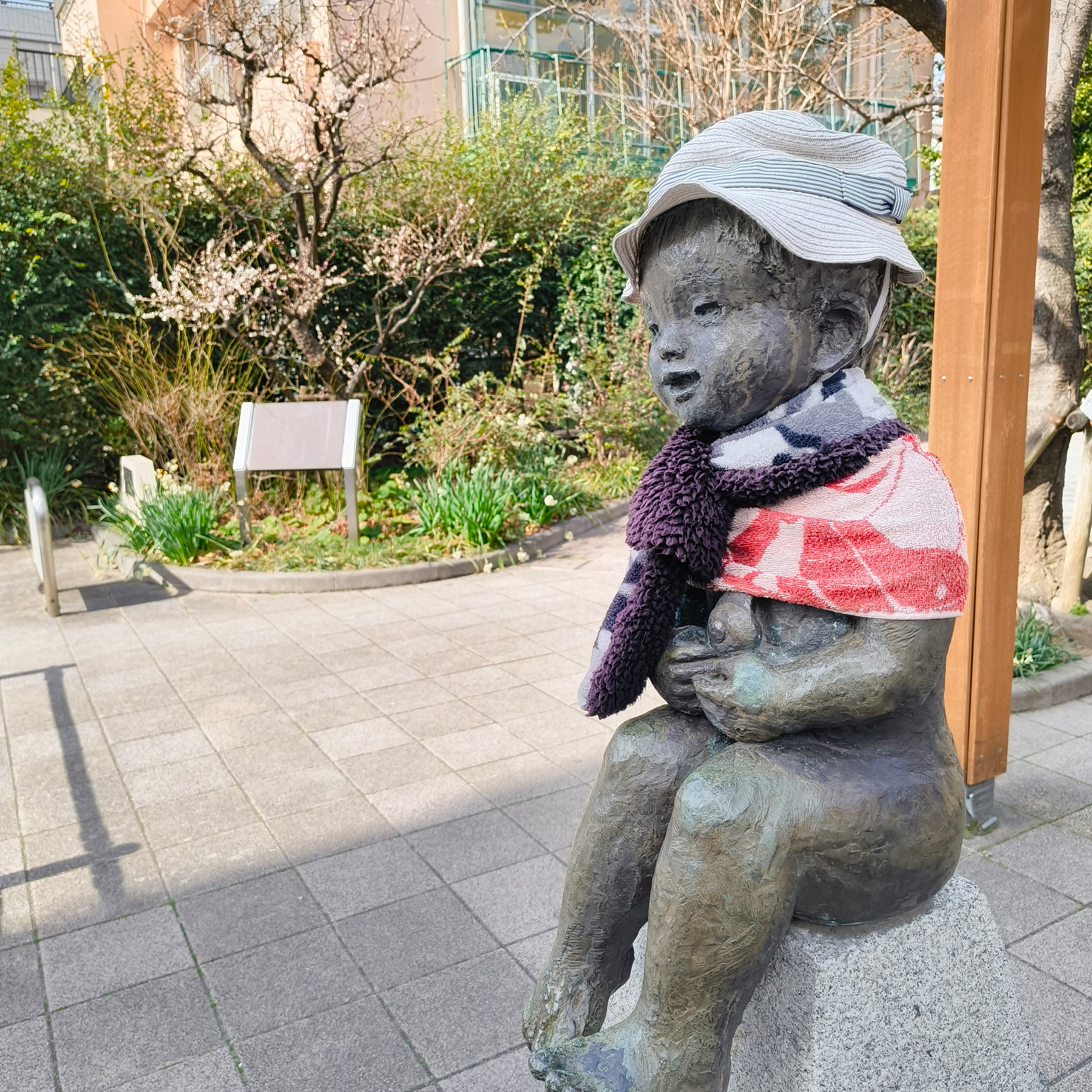 Auto-generated description: 公園の石の台に座っている毛糸の帽子とマフラーを身に着けた子供のブロンズ像です。