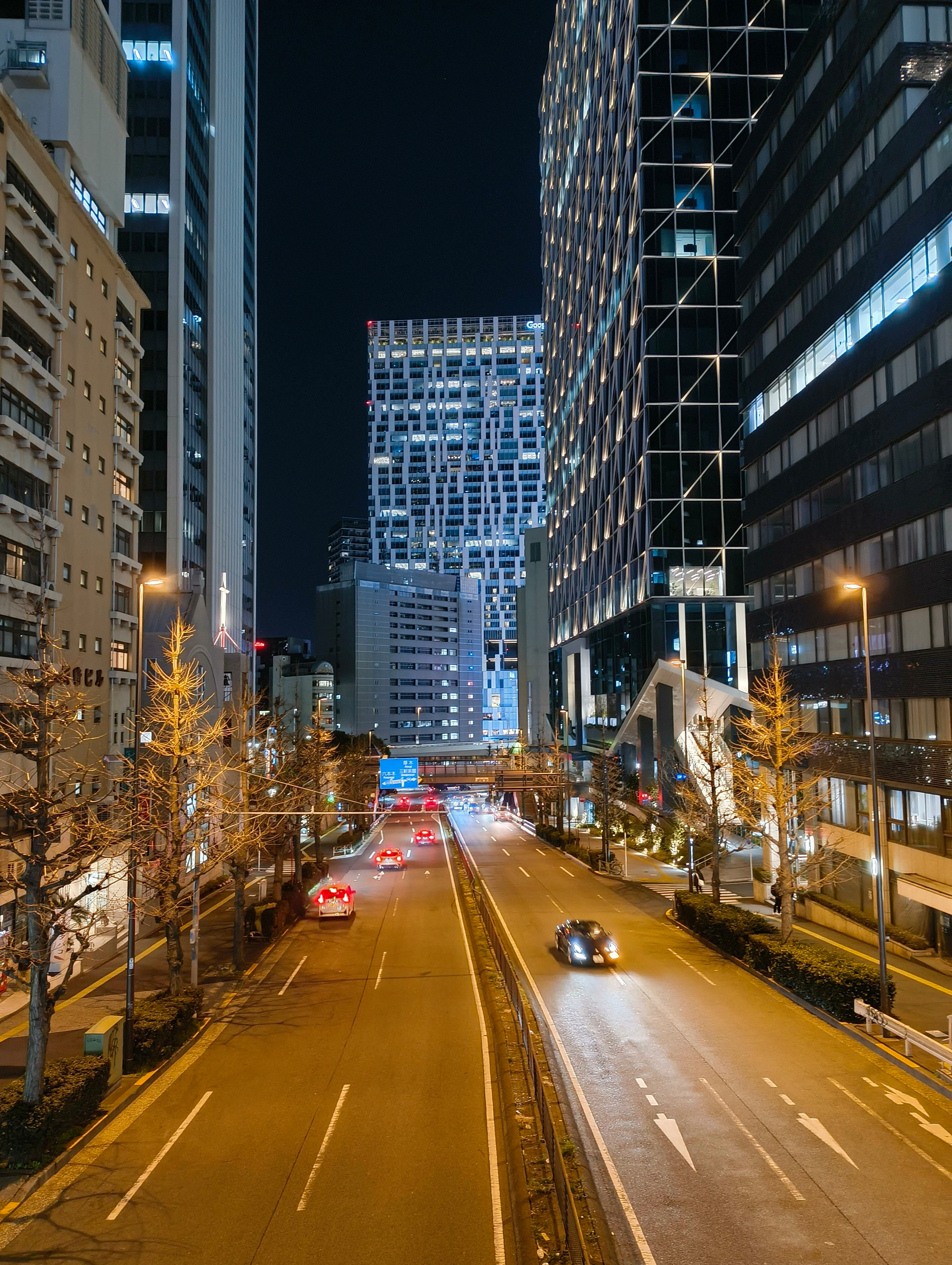 Auto-generated description: 高層ビルに囲まれた夜の市街地で、車が走行している道路が見える。