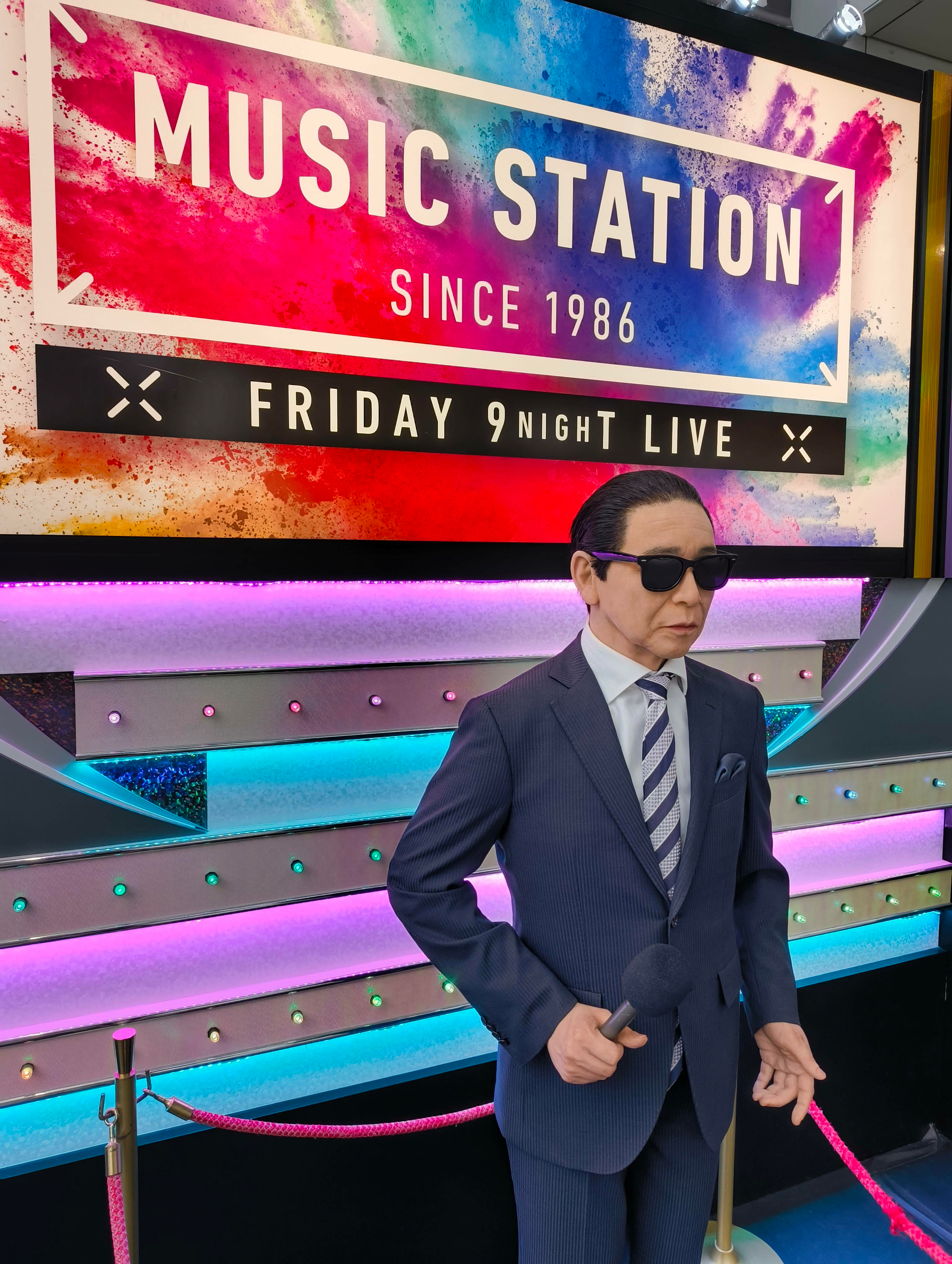 Auto-generated description: 色鮮やかな「MUSIC STATION」の看板の前で、タモリに似た蝋人形がマイクを持っている。