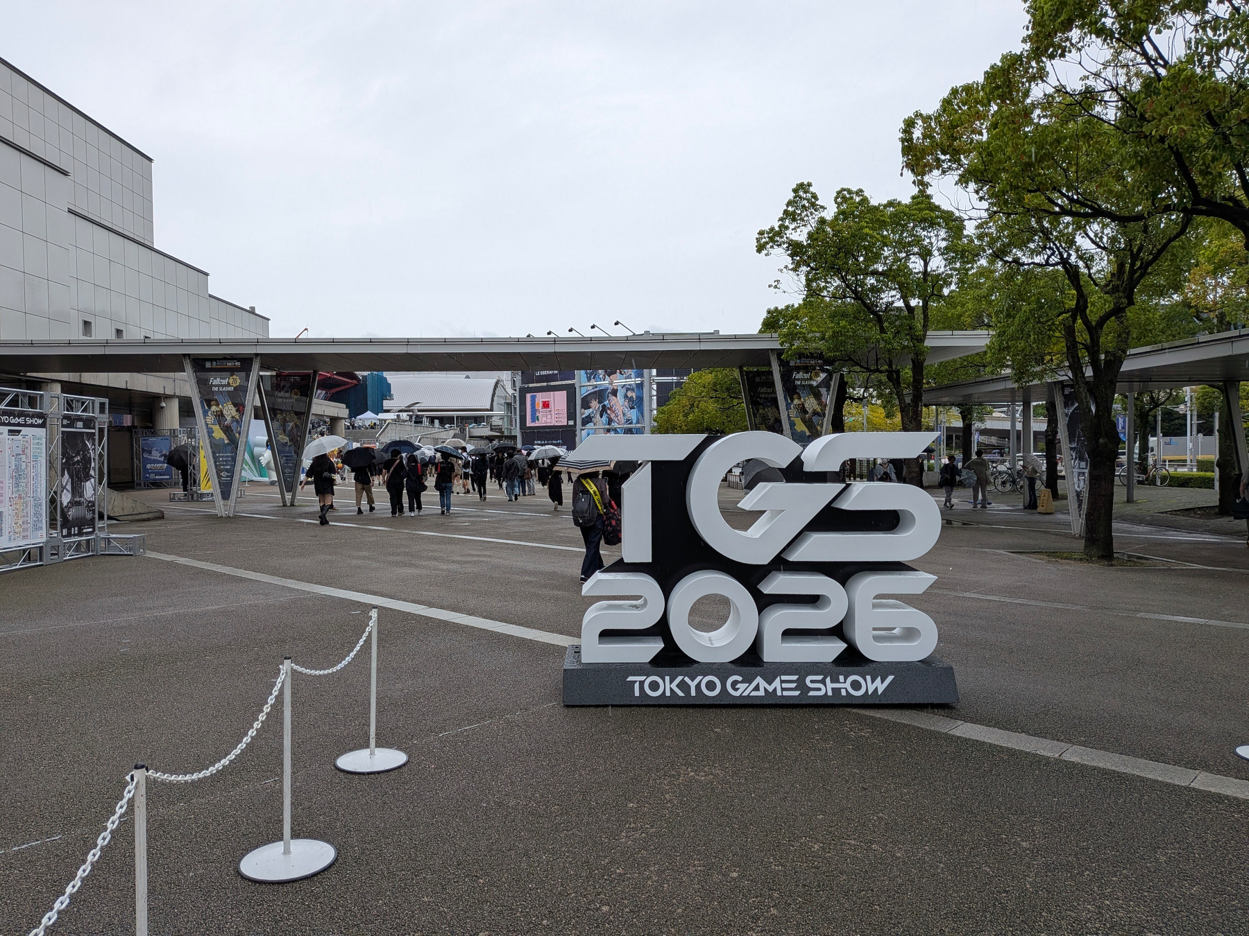 Auto-generated description: 2026年の東京ゲームショーのロゴが屋外に設置され、多くの人々が傘をさして訪れている場面。