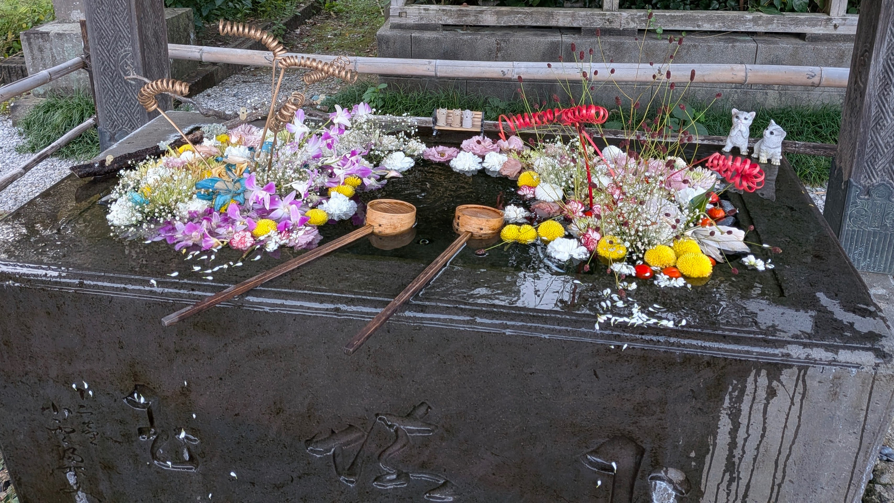 Auto-generated description: 花や装飾品が水をたたえた石の手水舎に美しく浮かべられている。